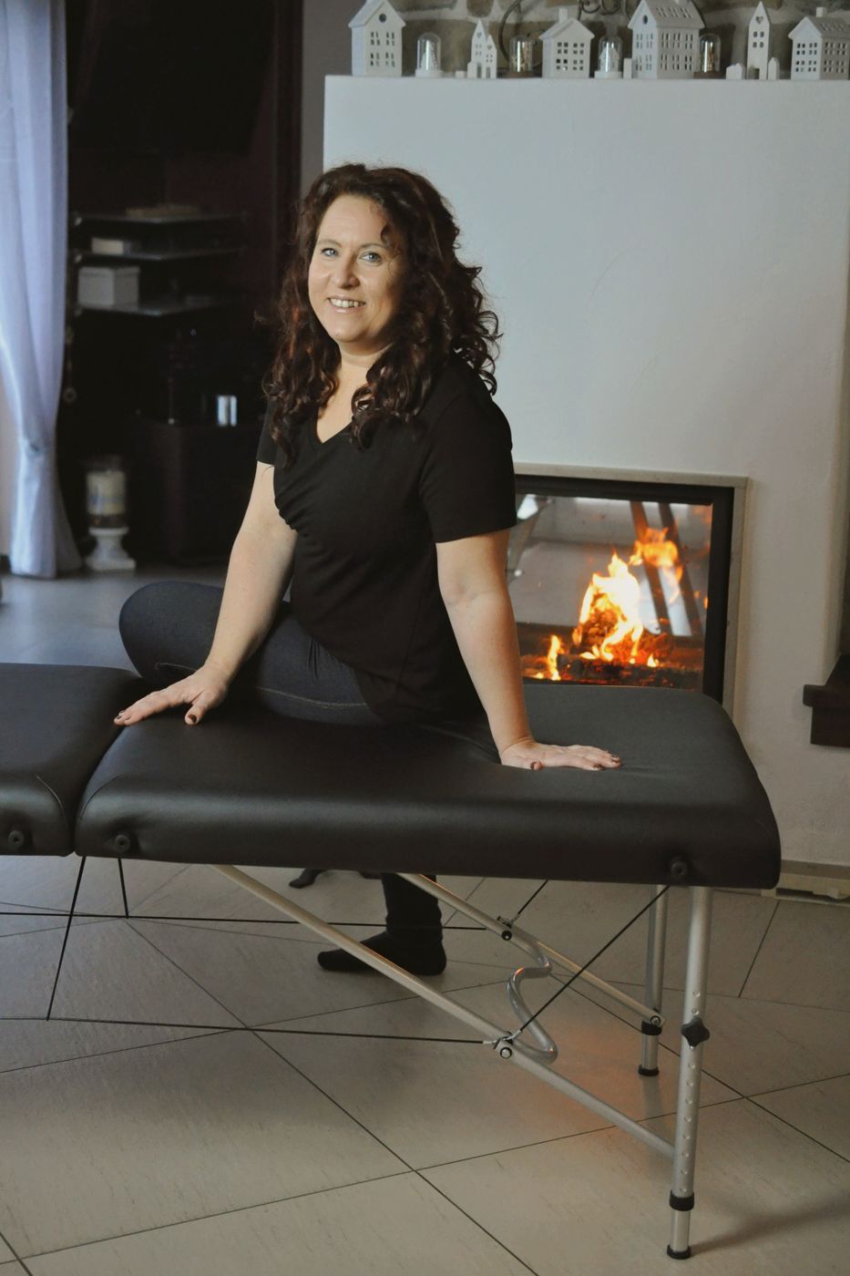 Manuela sitzt lächelnd auf ihrer Massageliege vor dem Kamin